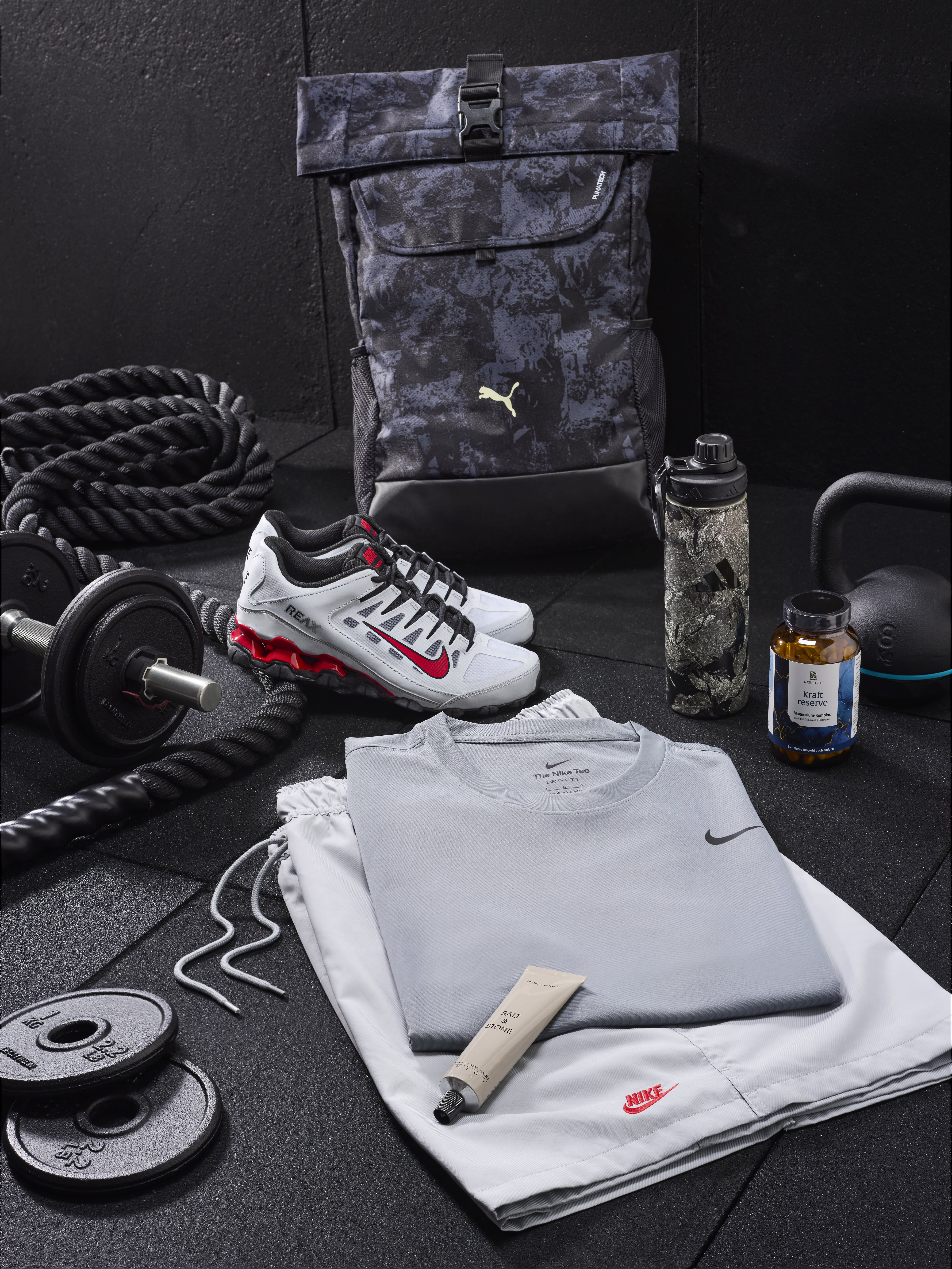In einer Gym-Kulisse sind Hanteln, Gewichtsscheiben, weiße Sportschuhe, ein dunkelgrauer Rucksack und hellgraue Sportklamotten zu sehen