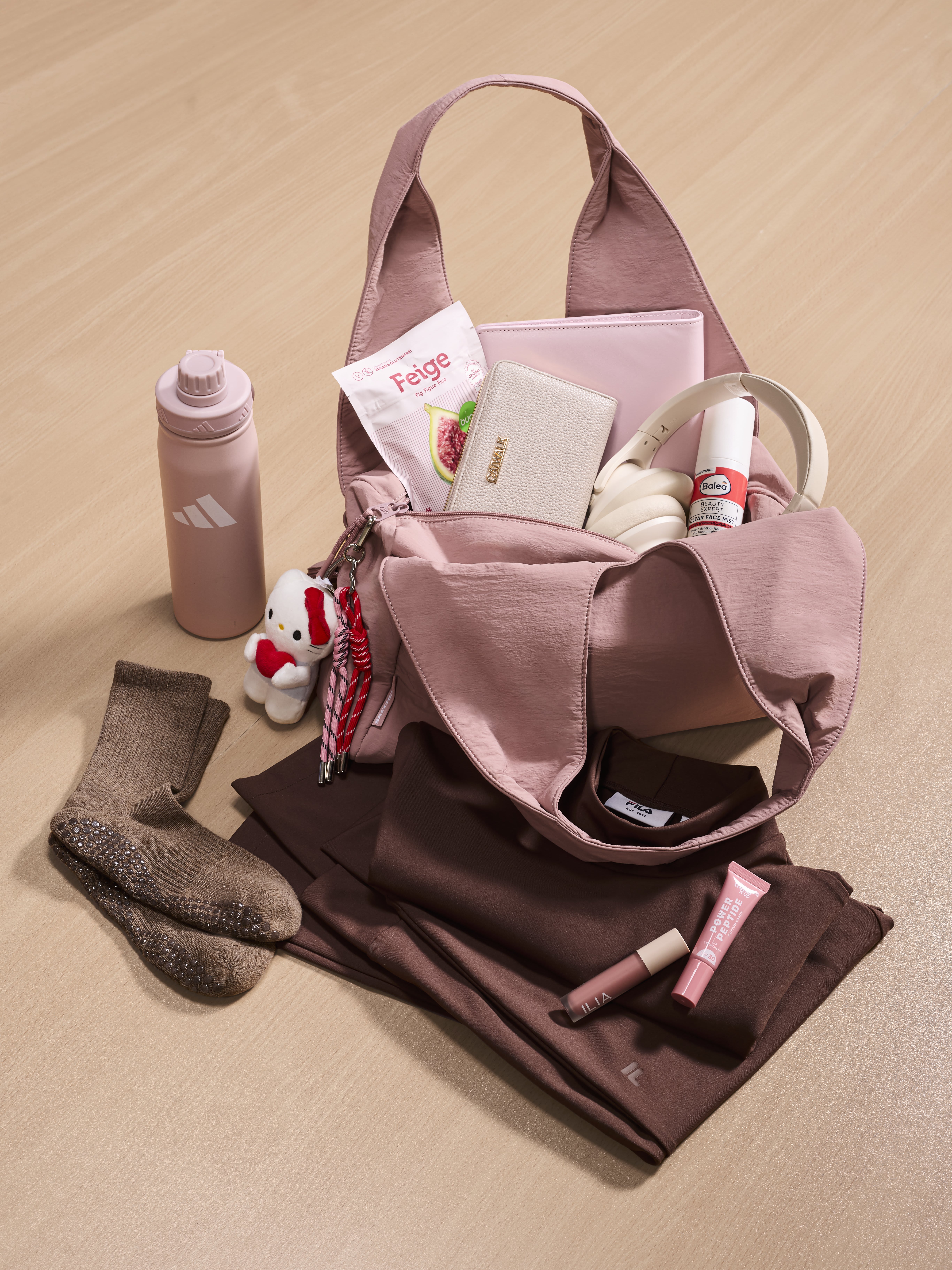 Eine rosafarbene Sporttasche mit Kopfhörern, Trinkflasche, Sportkleidung und kleinen Accessoires liegt auf einem hellem Untergrund