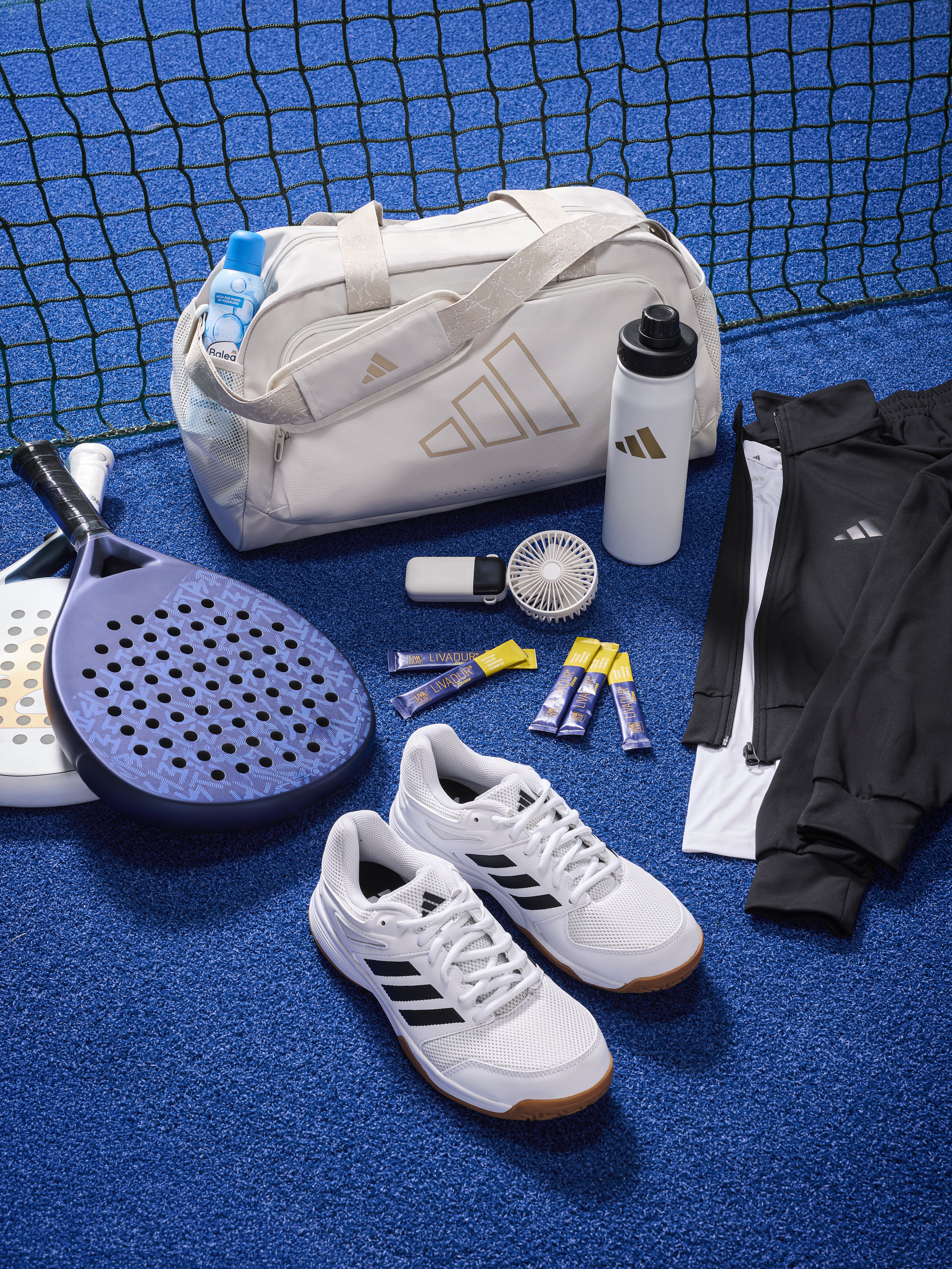 Hochformat: Padel-Schläger, eine Sporttasche, Sportschuhe, eine Trinkflasche und Sportklamotten stehen auf einem blauen Untergrund