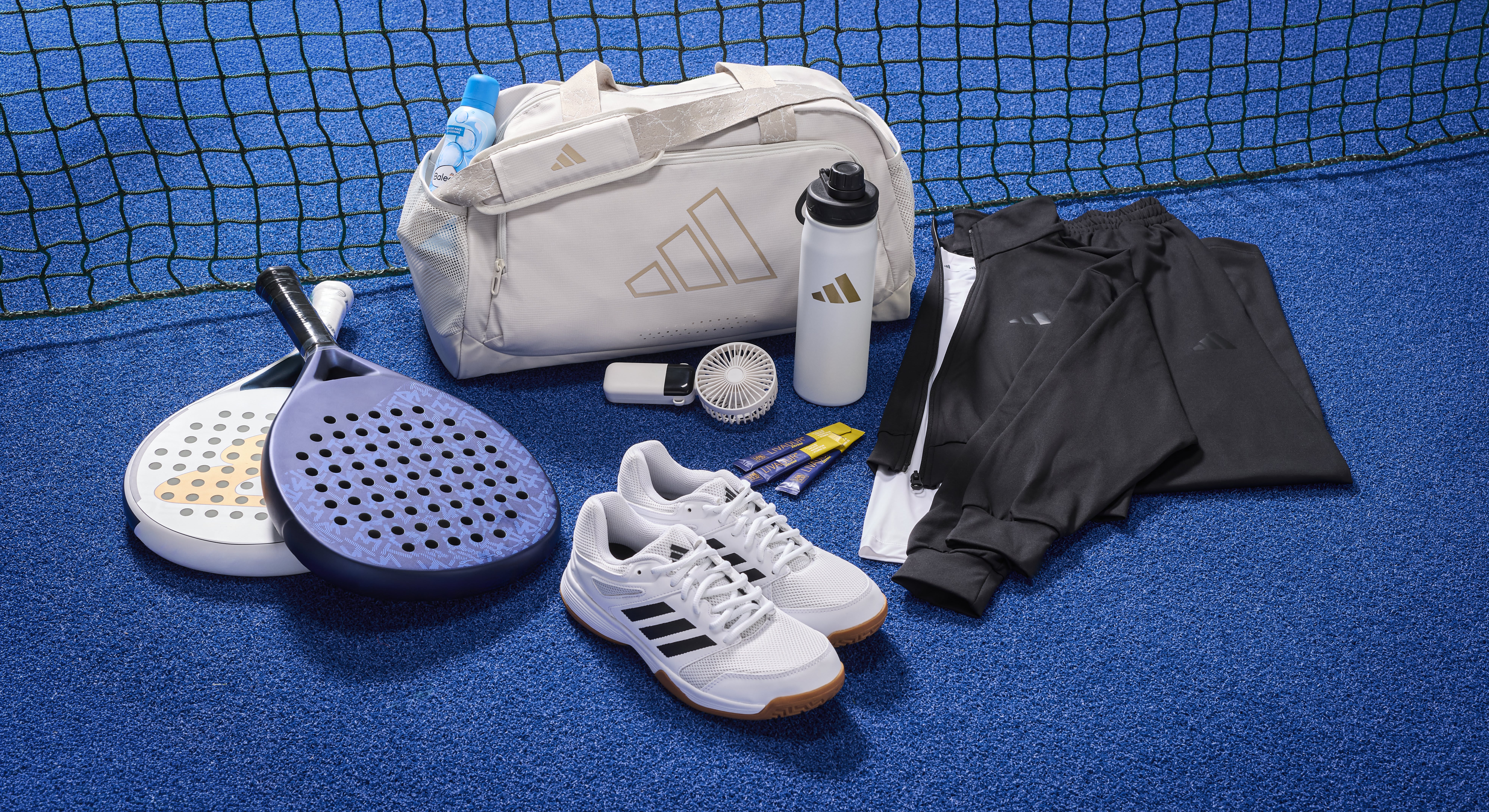 Padel-Schläger, eine Sporttasche, Sportschuhe, eine Trinkflasche und Sportklamotten stehen auf einem blauen Untergrund