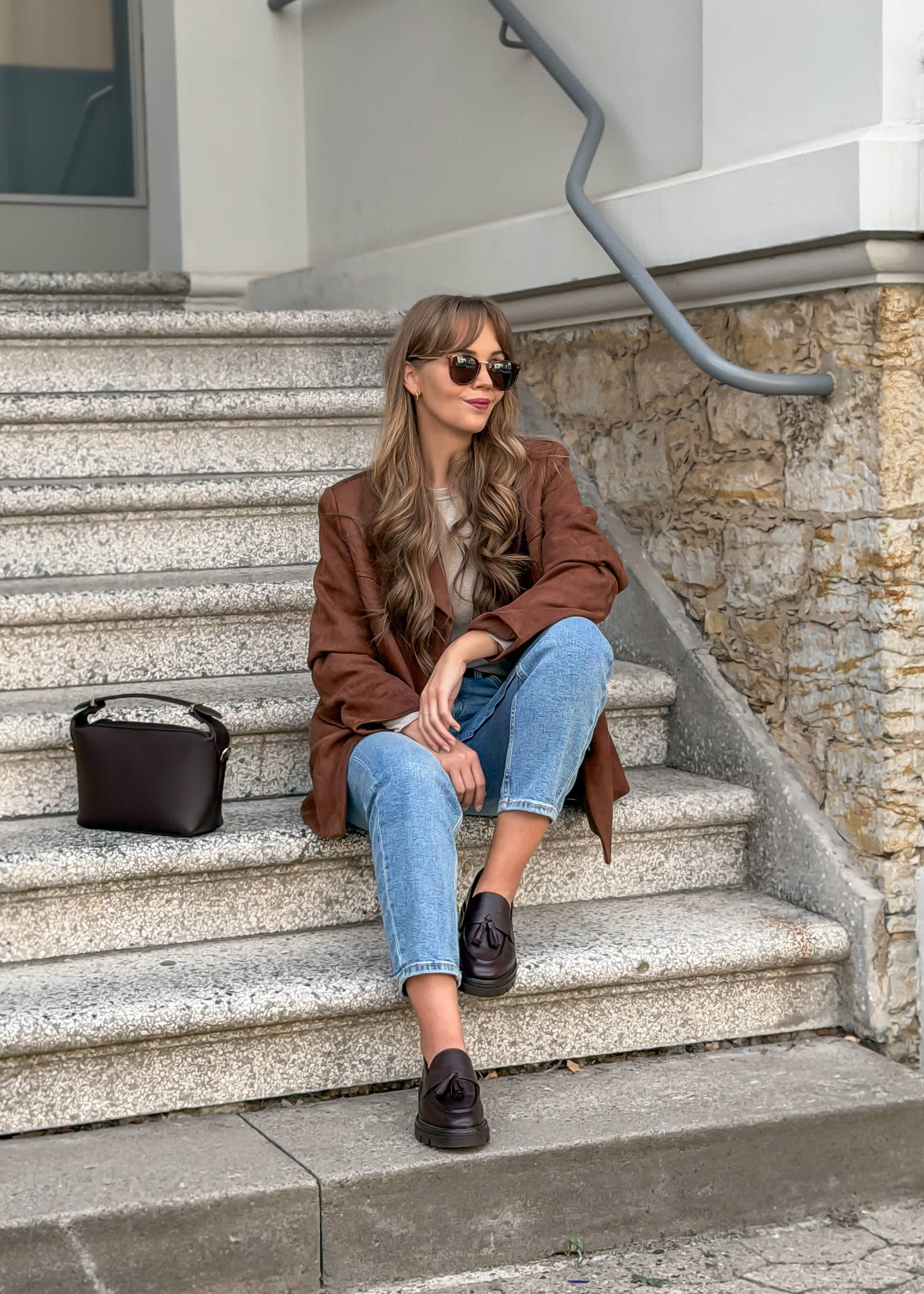 Eine Frau sitzt auf Treppe, trägt braunen Blazer, eine Jeans, dunkelbraune Loafer und passende Handtasche