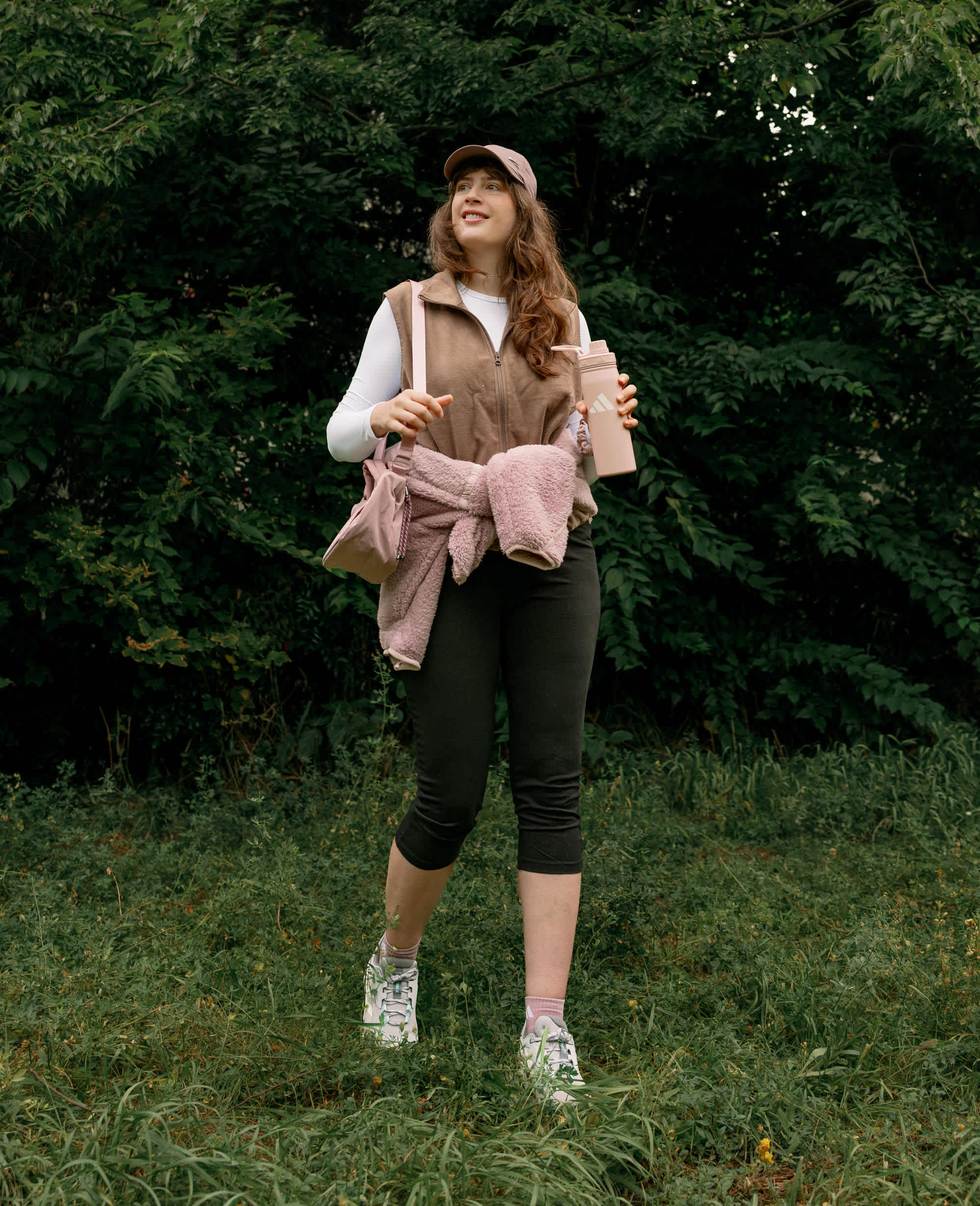 Frau in Wanderkleidung mit rosa Fleecejacke, Weste, Capri-Leggings und Cap im Grünen