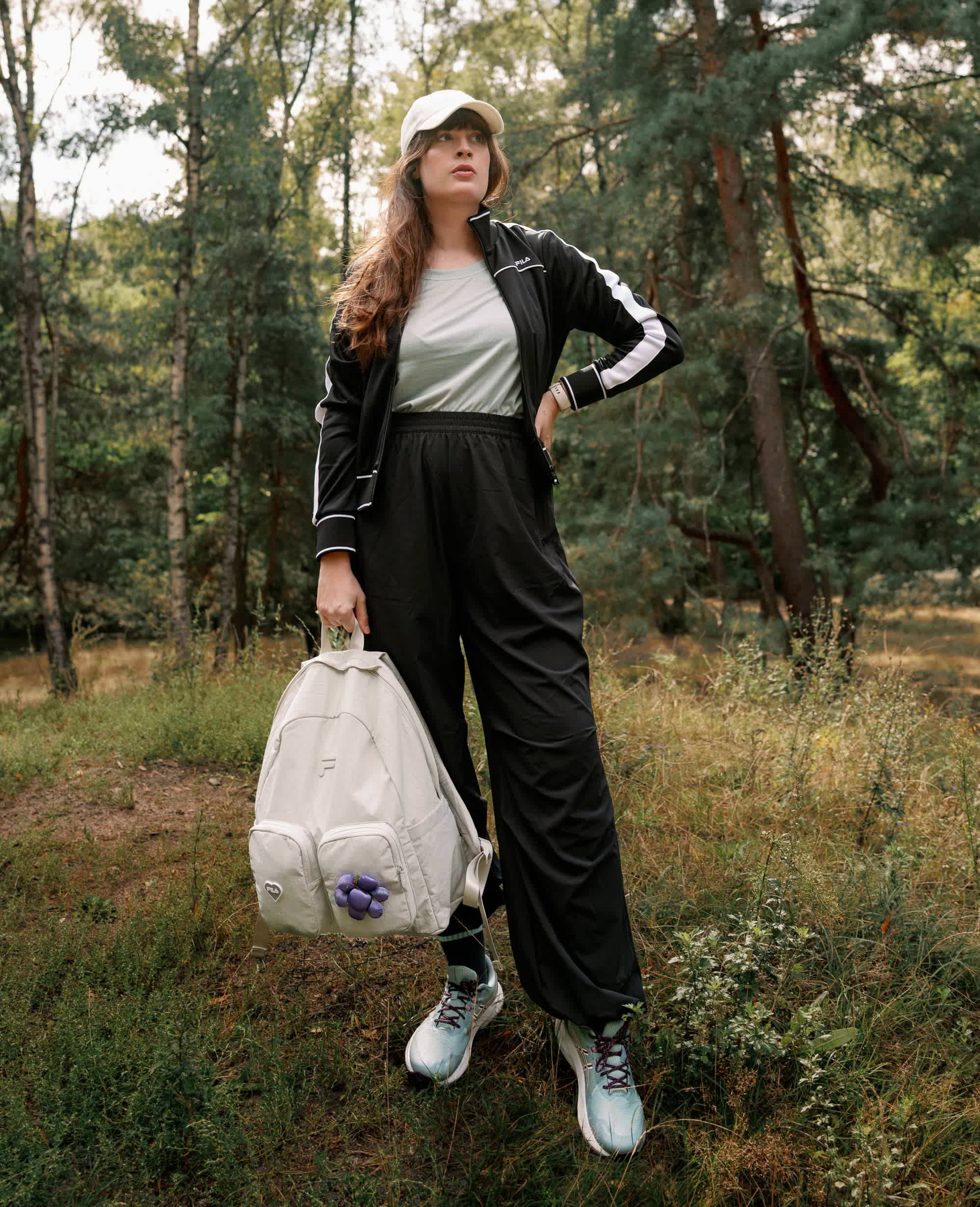 Frau in sportlichem Outfit mit weiter schwarzer Hose, Jacke und Sneakern steht mit einem Rucksack in einem Wald