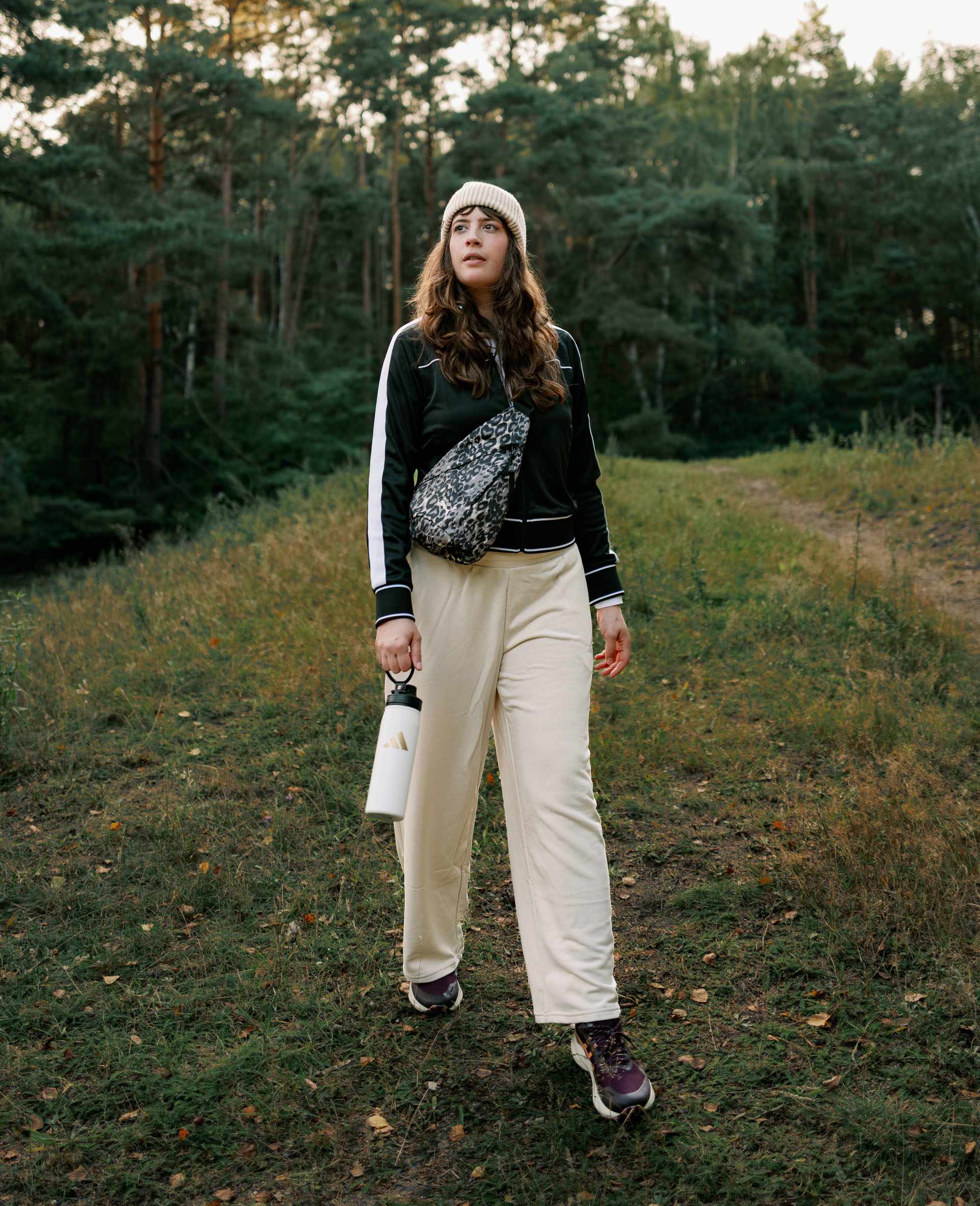 Frau in schwarzer Trainingsjacke und beiger Hose steht mit Leopard-Crossbody-Bag und weißer Trinkflasche auf einem Waldweg