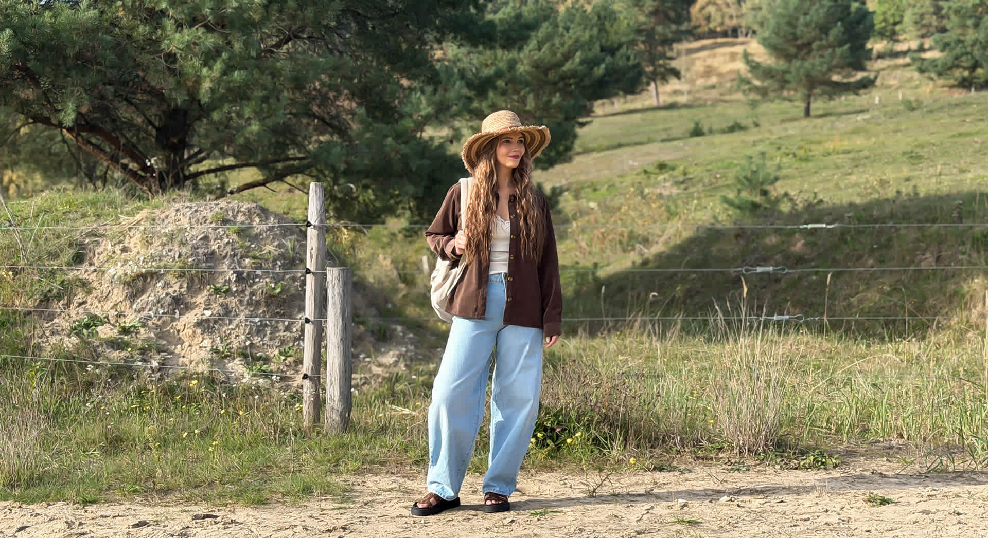 Frau mit Strohhut in brauner Cordjacke und Wide-Leg-Jeans trägt Plateau-Sandalen auf einem Feldweg