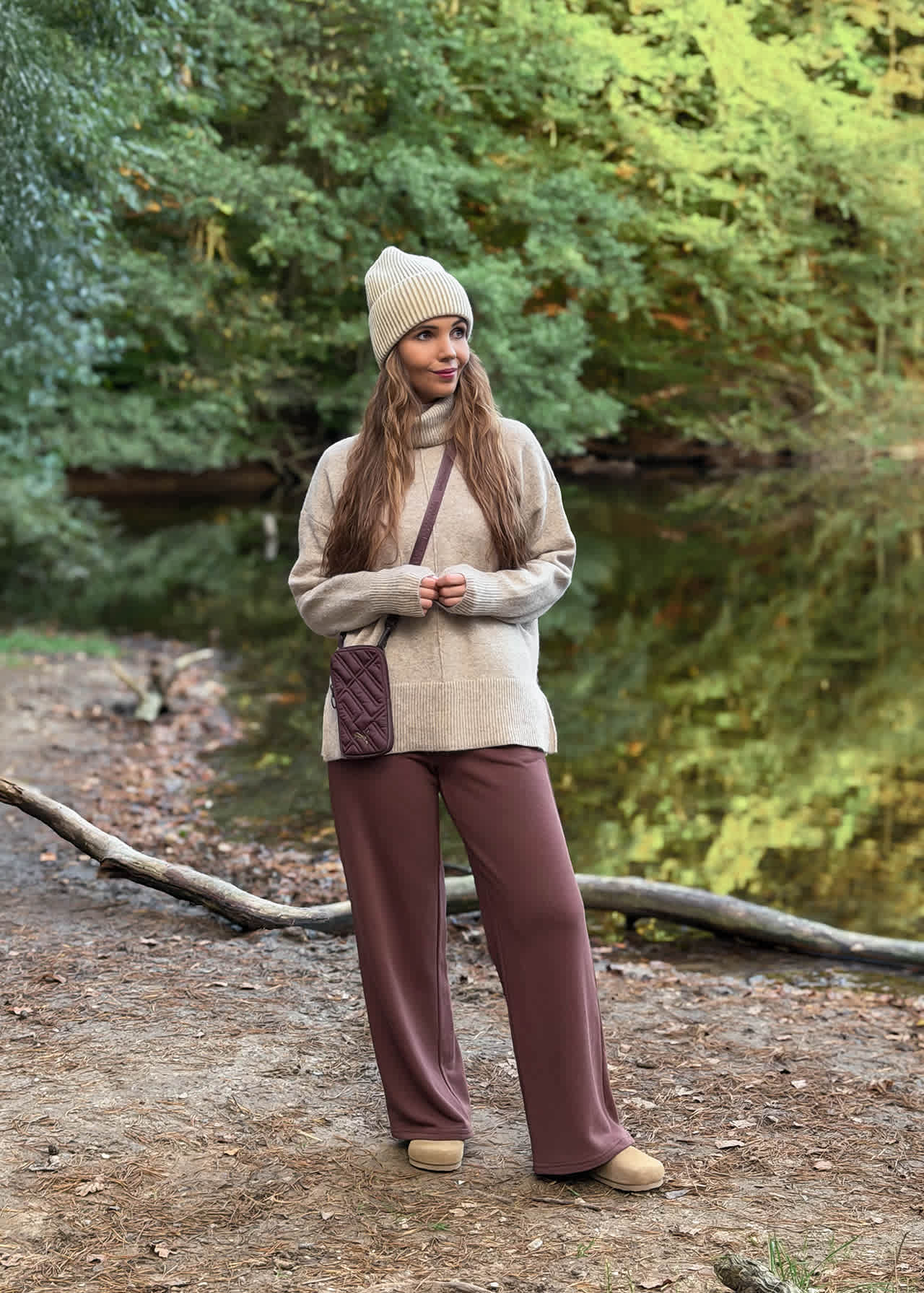 Frau in beigem Rollkragenpullover, Beanie und brauner Wide-Leg-Hose trägt Plateau-Boots am Waldsee