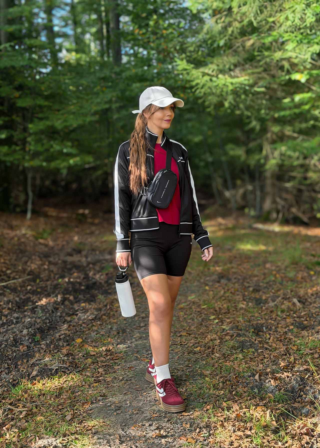 Frau in Glamping Outfit mit Trackjacke, Radlerhose und Cap trägt bordeauxrote Sneaker im Wald