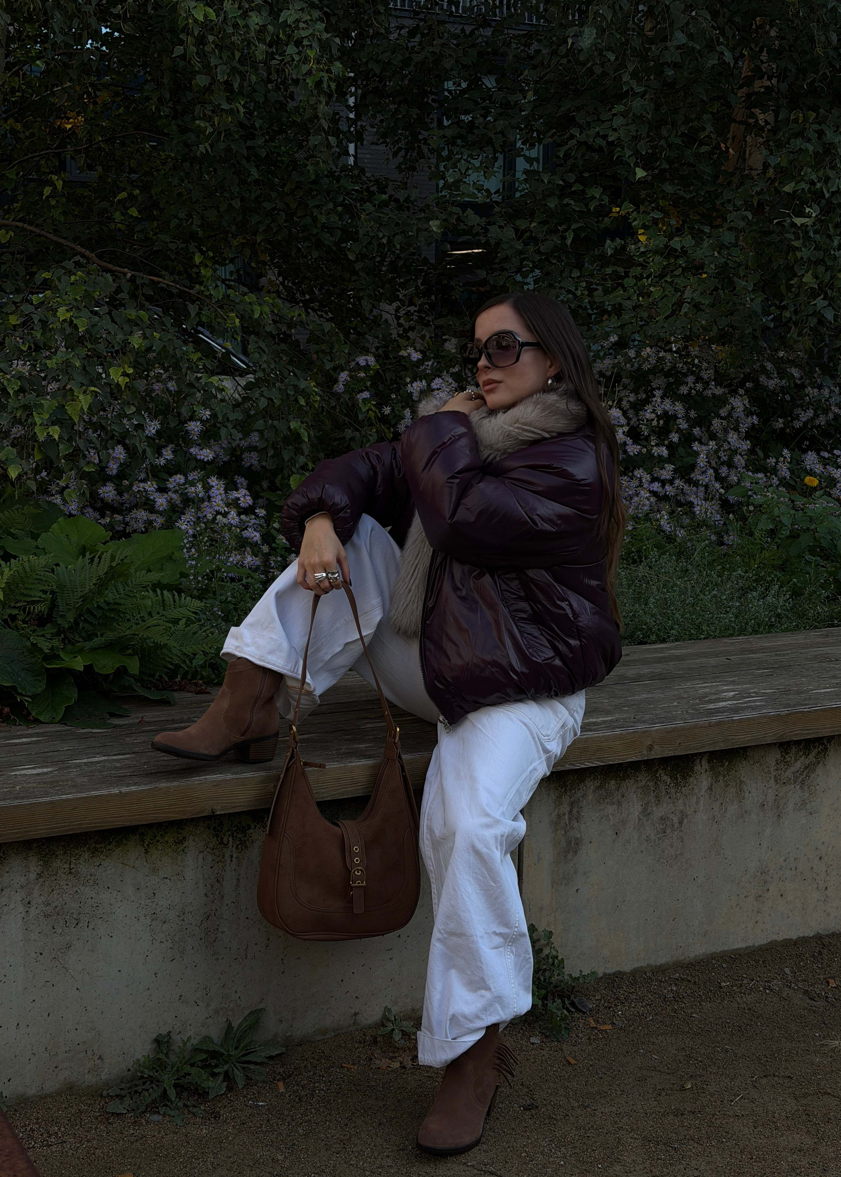 Frau auf Holzbank in Daunenjacke mit Fellkragen und weißer Jeans trägt braune Westernstiefeletten und Boho-Bag