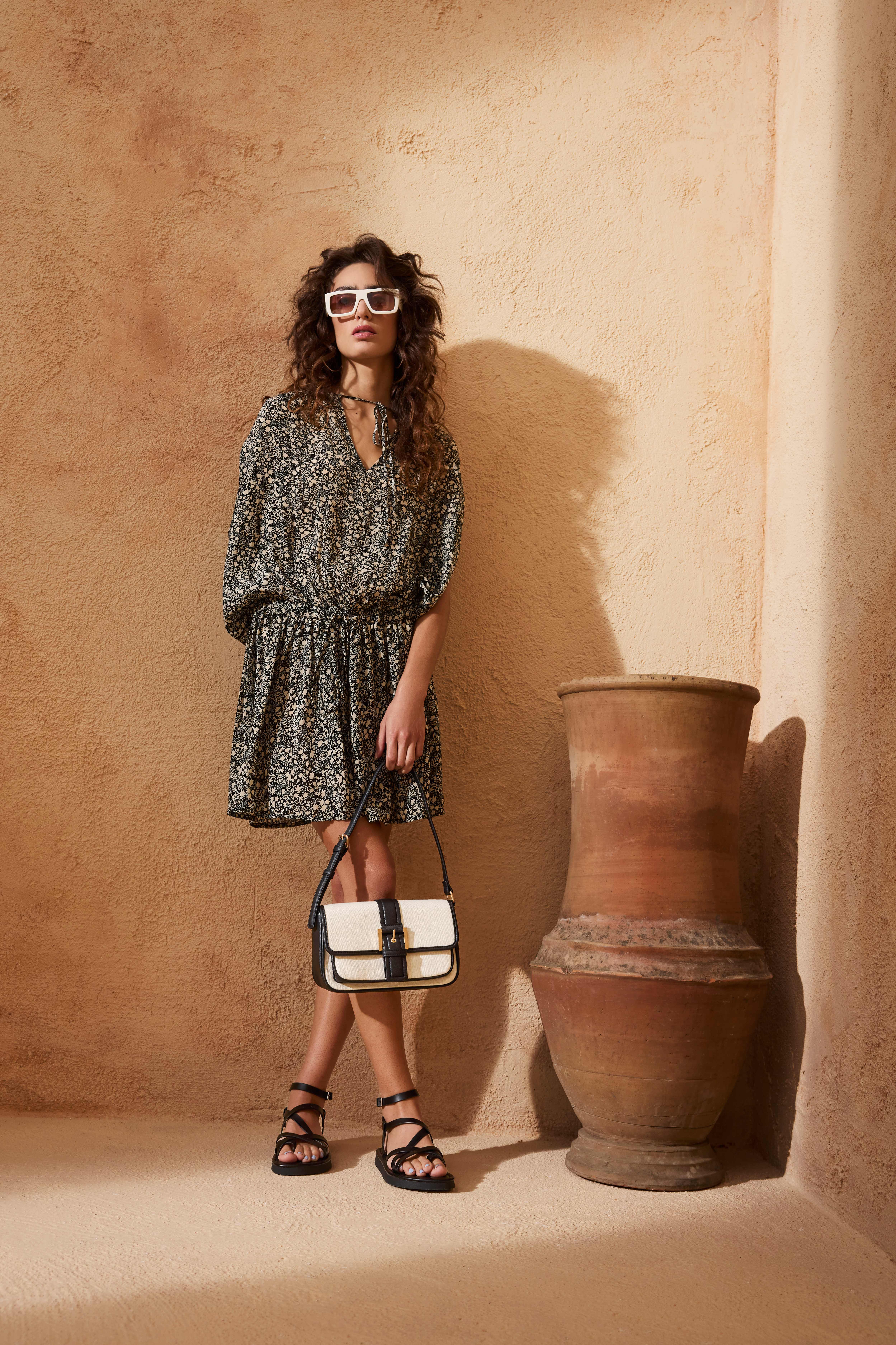 Frau in gepunktetem Kleid und weißer Sonnenbrille, steht mit überkreuzten Beinen und offenen Sandalen vor Backstein-Wand