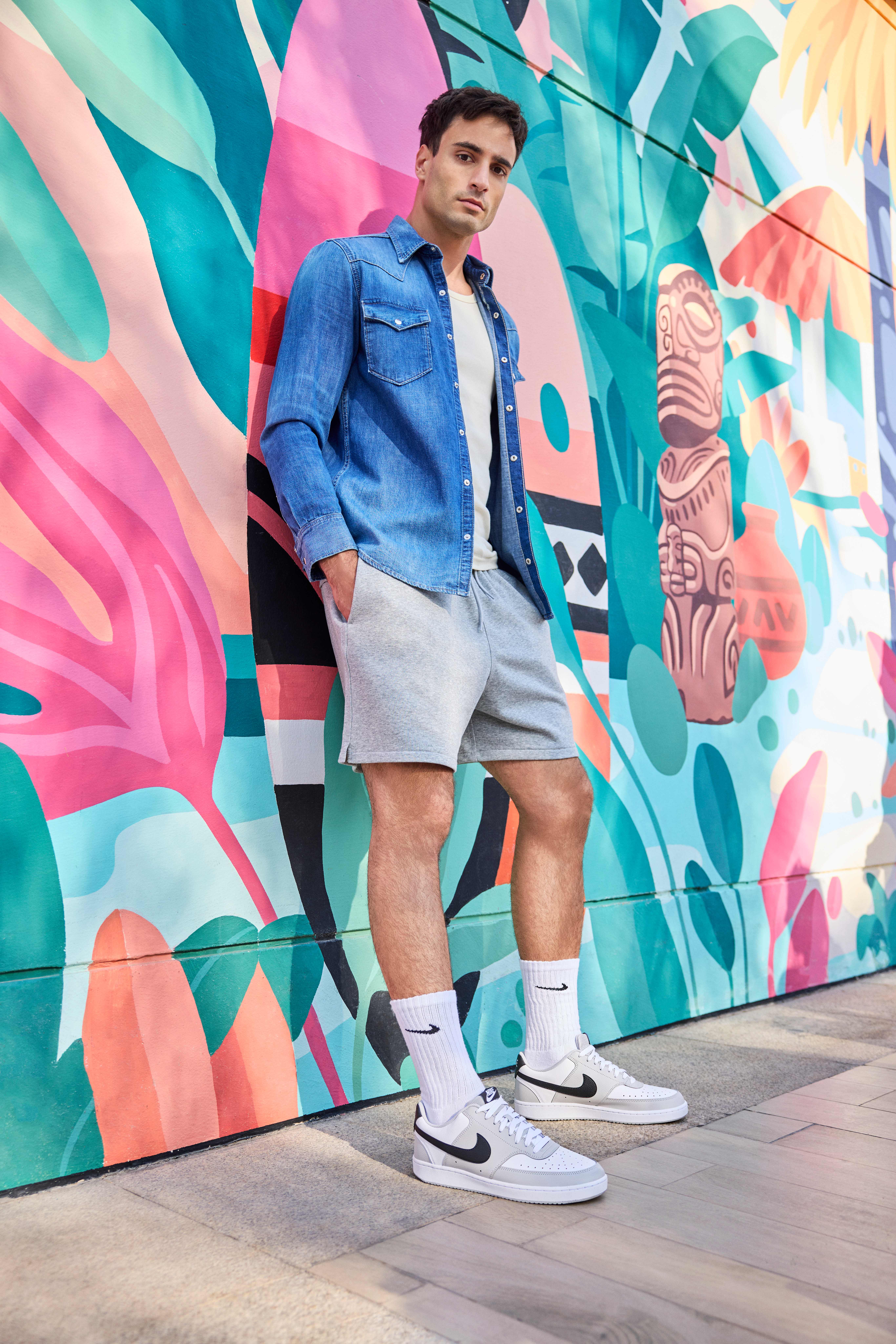 Mann lehnt lässig an bunter Wand, trägt Denim-Hemd, kombiniert mit weißem Shirt, grauen Shorts und weißen Nike-Sneakern
