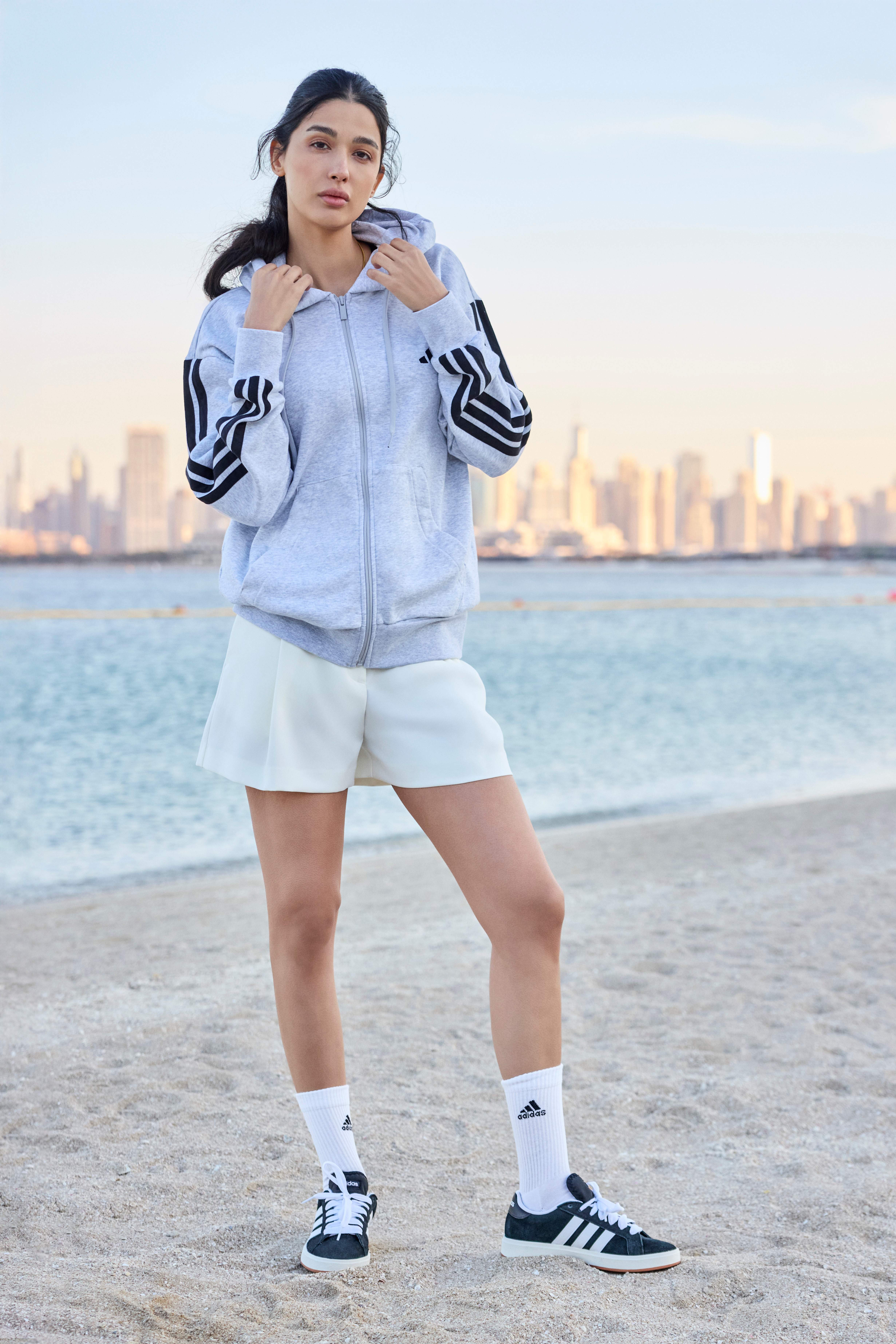 Frau trägt sportliches Outfit, bestehend aus Sport-Sweater und Faltenrock, stehend mit grünen Sneakern am Strand