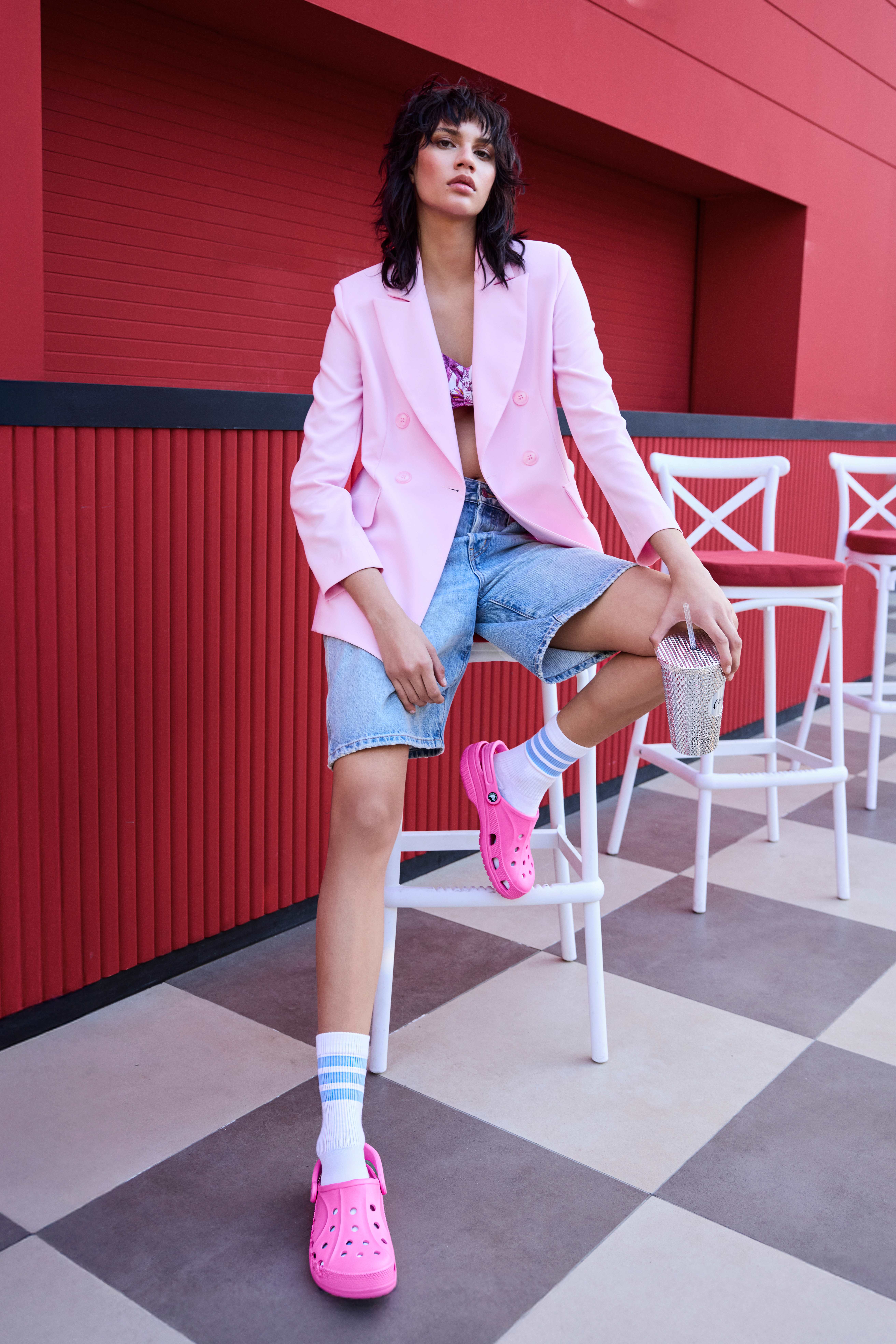 Frau sitzt entspannt auf Barhocker, gekleidet in rosa Blazer und blaue Shorts, mit pinken Crocs