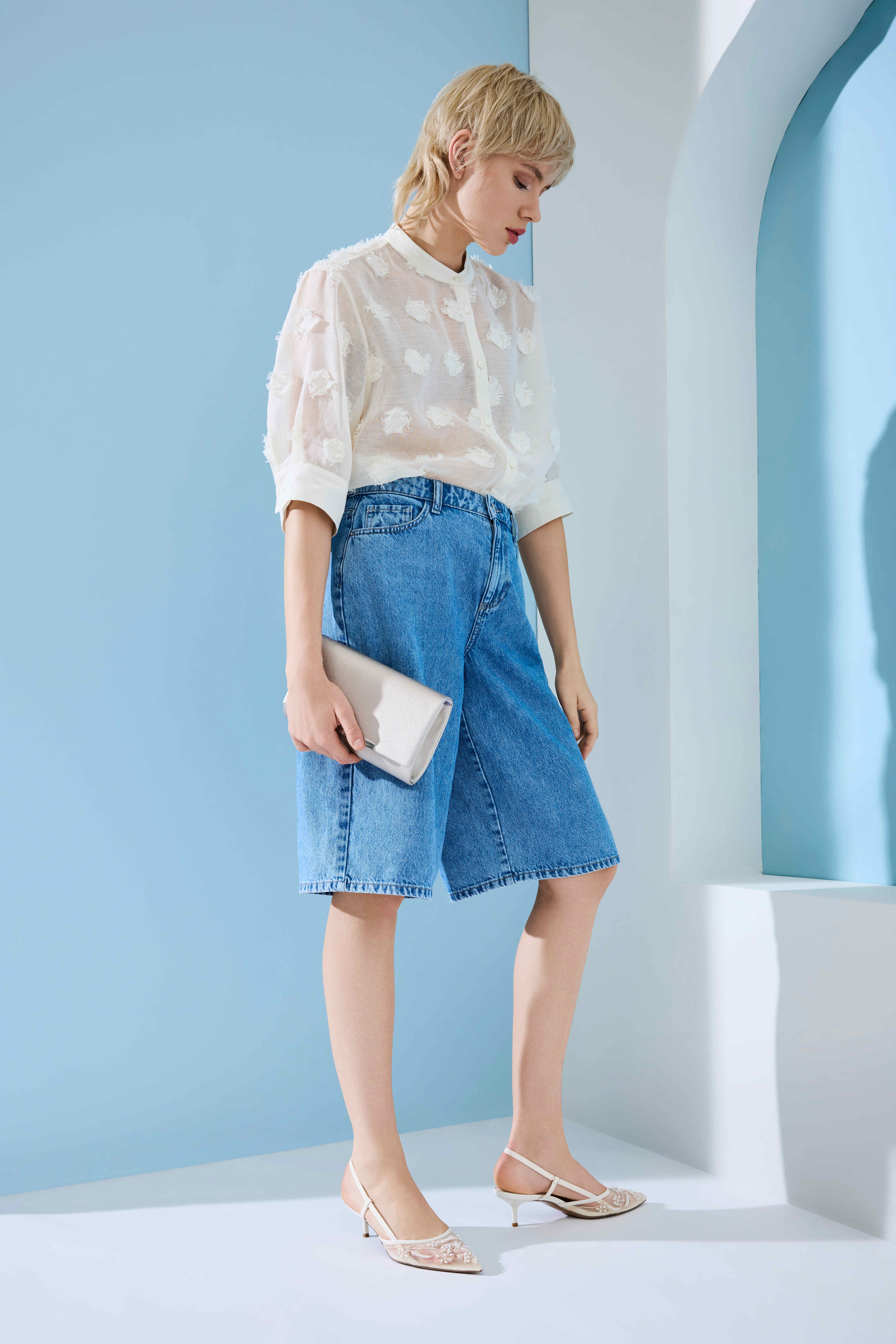 Kurzhaarige blonde Frau mit transparentem Top, hohen Denm-Shorts und Slingbacks