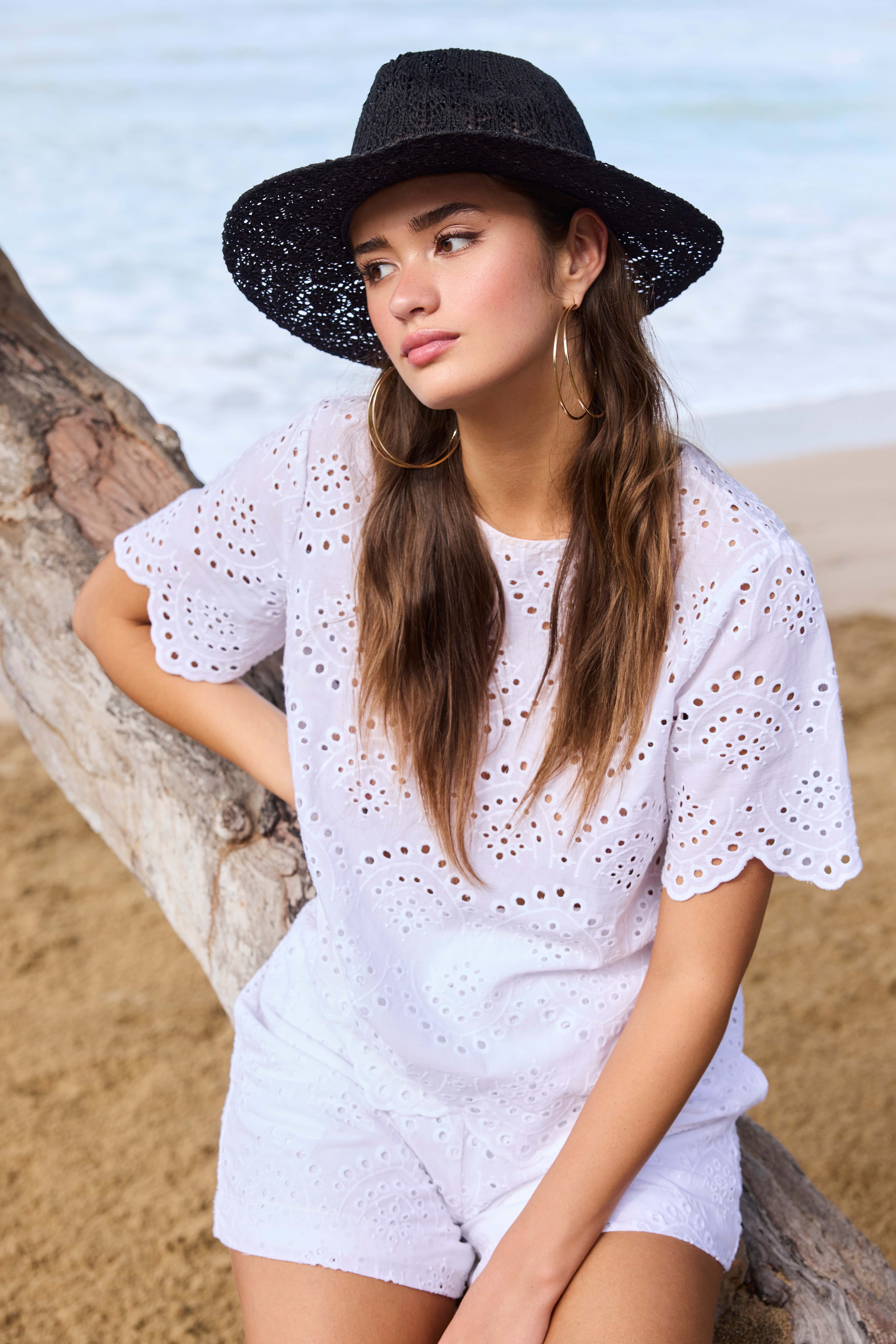 Frau mit schwarzem Sommerhut und weißem Spitzenkleid, sitzt auf Baumstamm am Strand