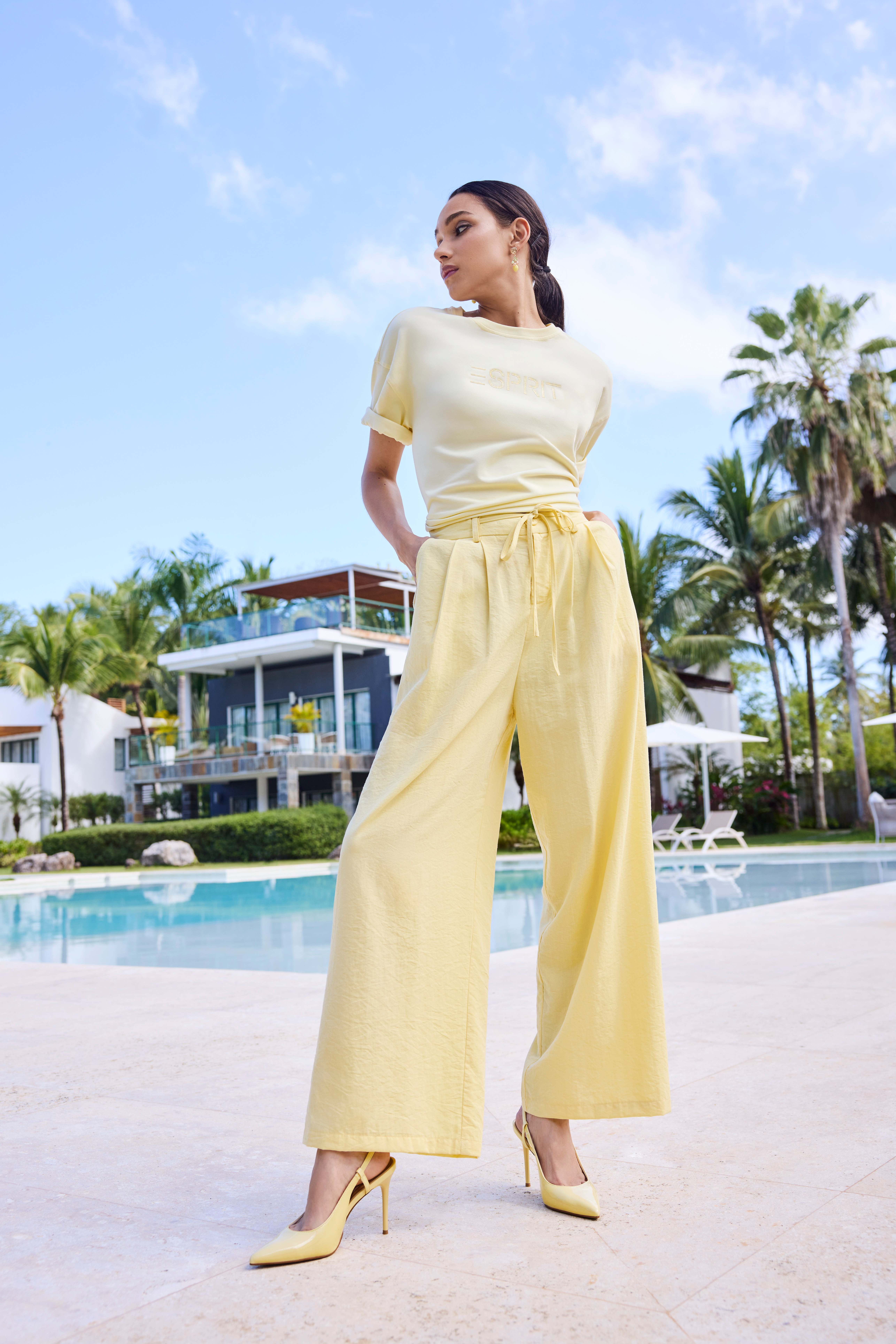 Frau in hellgelbem Outfit, mit passenden Pumps, steht vor Pool