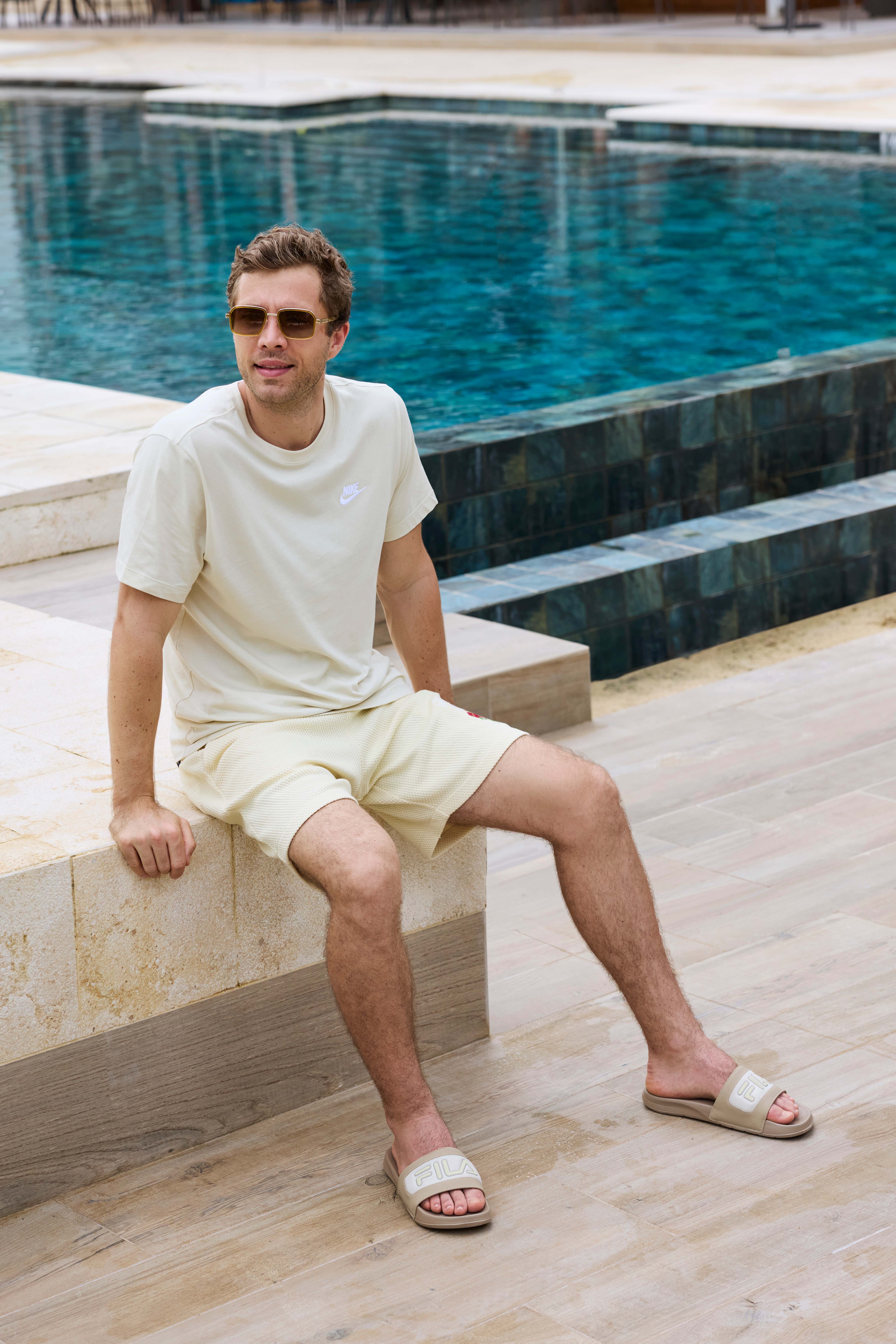 Mann in sandfarbenem Shirt und passenden Shorts, trägt Sonnenbrille, sitzt am Pool