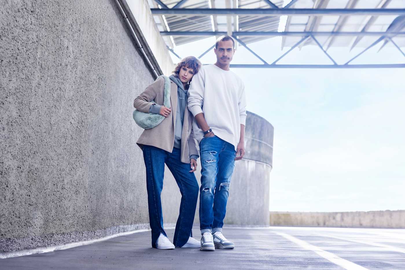 Frau und Mann stehen lässig nebeneinander, gekleidet in Alltagskleidung mit Jeans, Sweatern und Sneakern, mit Blick zur Kamera