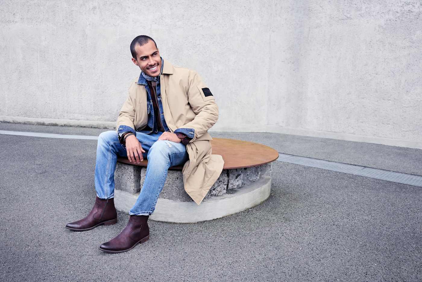 Mann sitzt auf Bank, trägt beige Jacke, Jeanshose und Cowboy Boots