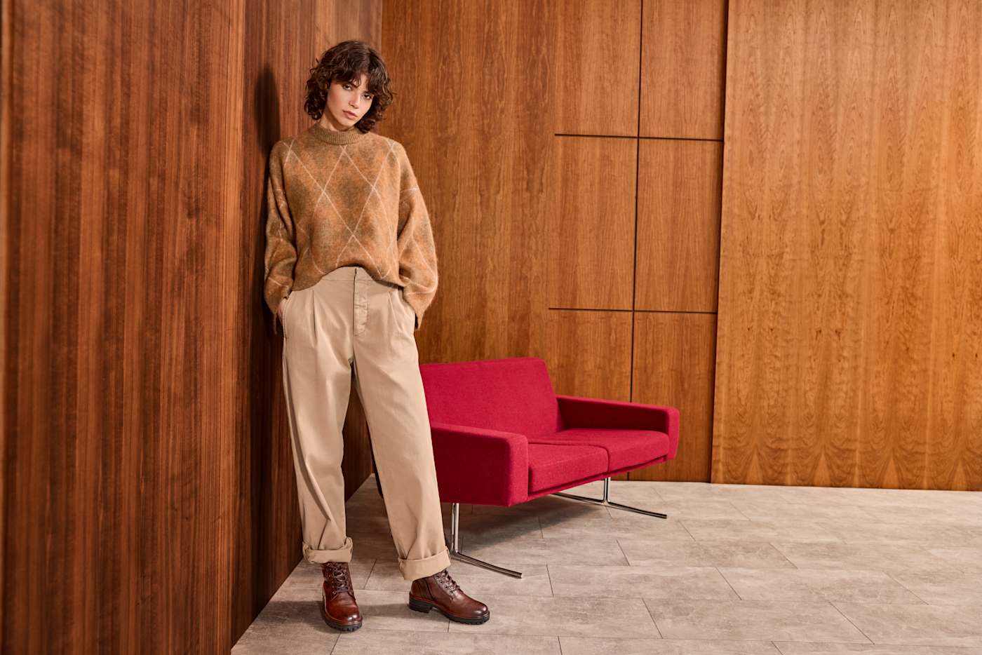 Frau in braunem Wollpullover und gemütlicher Palazzohose, lehnt an Holzwand