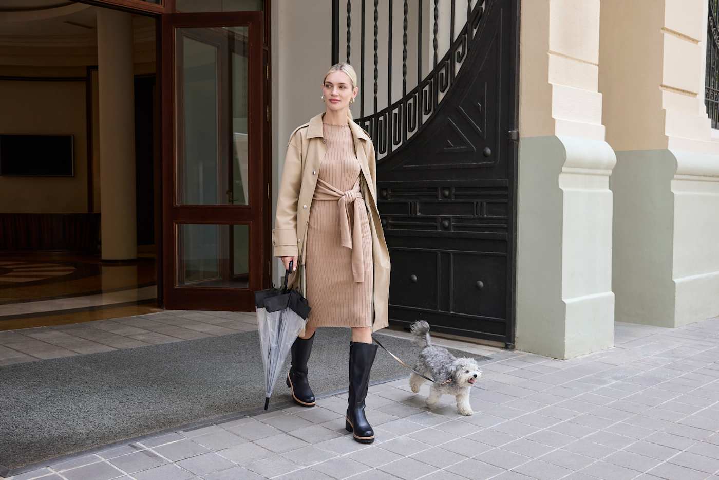 Frau in braunem Trenchcoat und schwarzen Stiefeln geht Gassi mit Hund