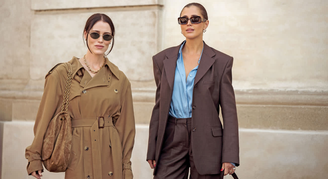 Zwei Frauen in schlichten Outfits, dem Copenhagen Streetstyle nachempfunden, links mit beigem Trenchcoat und passender Tasche, rechts mit braunem Oversize-Anzug und blauer Bluse