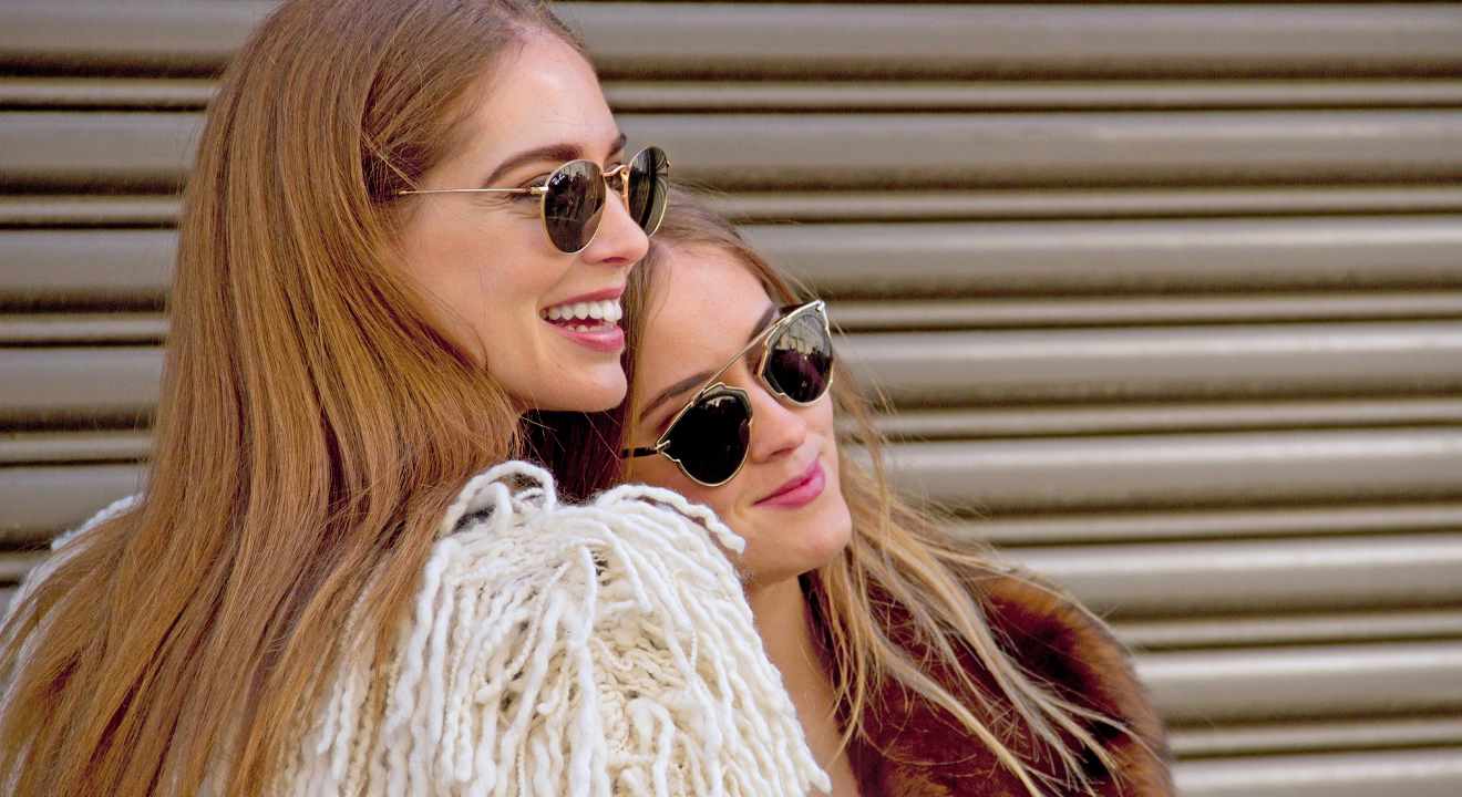 Zwei junge Frauen in Nahaufnahme, aneinander lehnend, mit Sonnenbrille und selbstsicherem Lächeln im Gesicht.