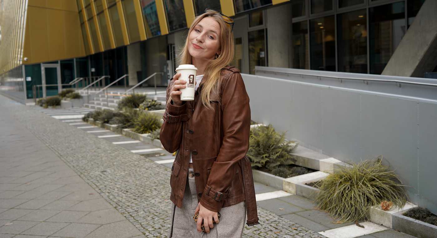 Frau in brauner Lederjacke mit Coffee-to-go-Becher, Outfit im Matcha Latte Mode-Stil, stehend vor Gebäude.