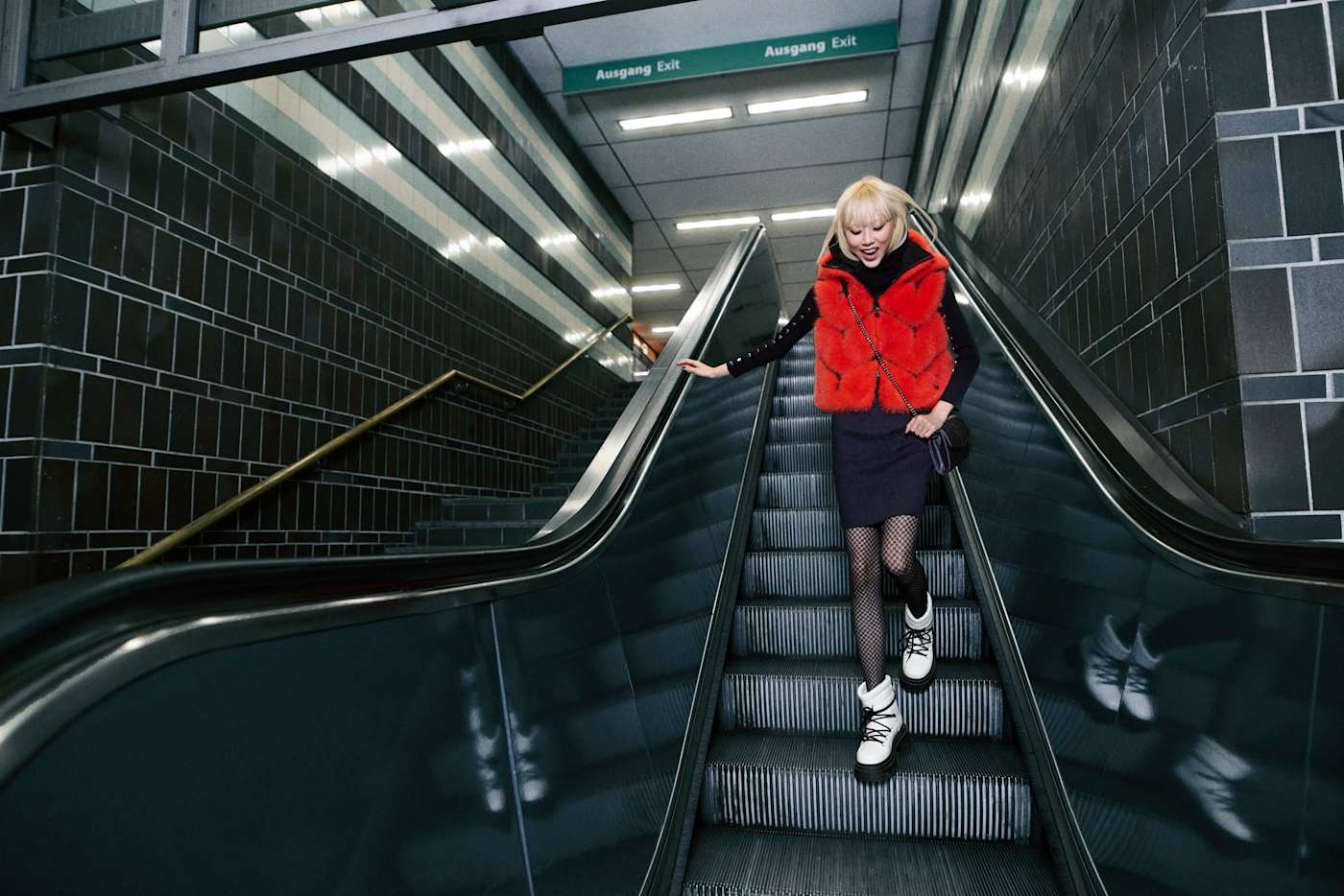 Frau mit roter Weste, schwarzer Netzstrumpfhose und weißen Schnürstiefeln auf einer Rolltreppe in einer U-Bahn-Station