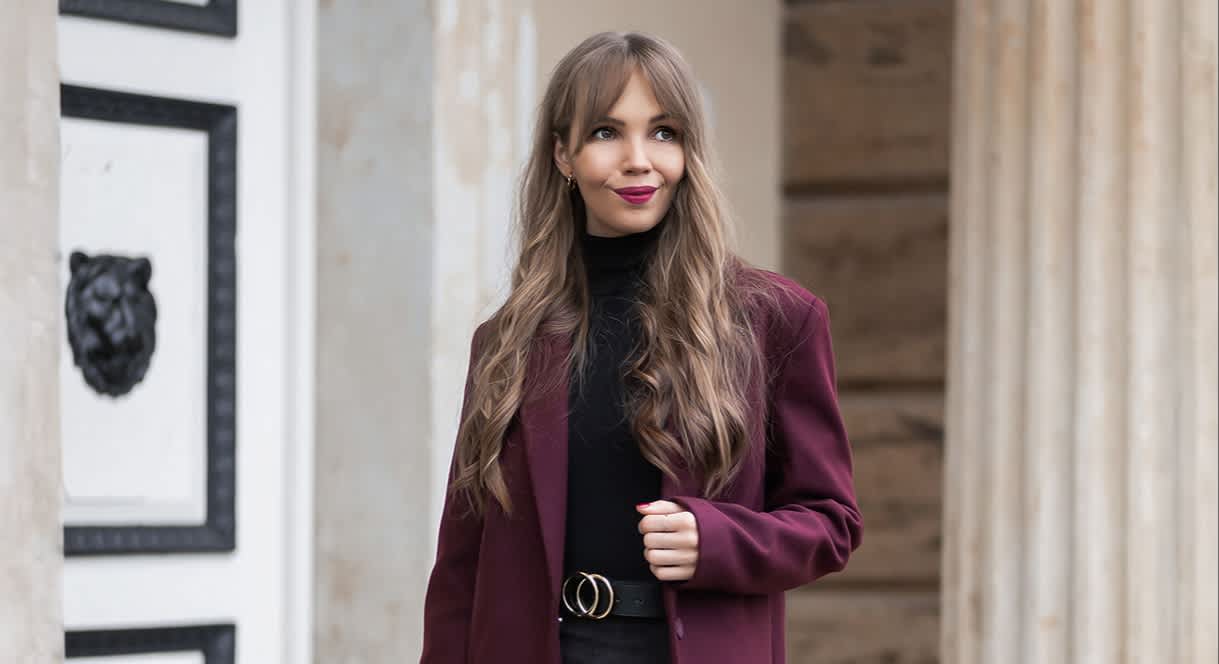 Frau mit burgunderfarbenem Mantel in Kombination mit schwarzem Pullover, Rock und Stiefeln, mit schwarzer Tasche in der Hand