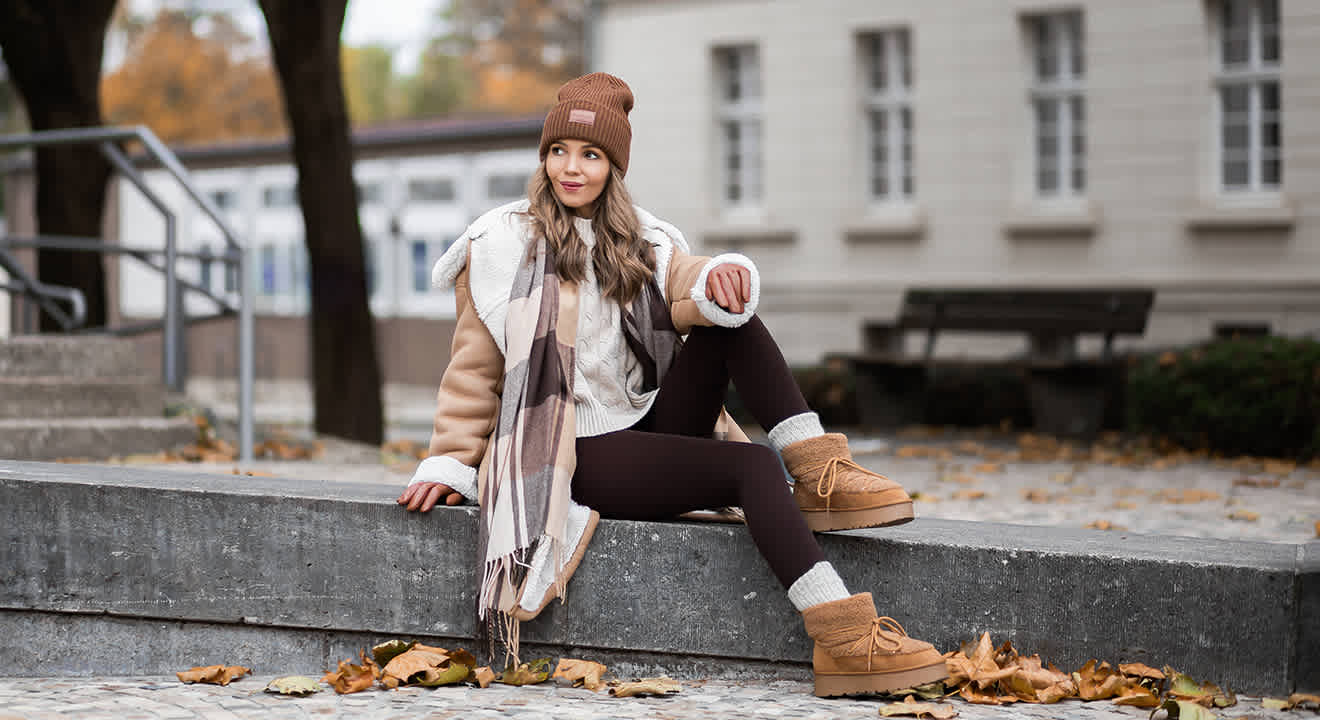 Frau in cosy Weihnachtsmarkt-Outfit mit brauner Mütze, Schal und warmen Fell-Boots, sitzend auf Steinfläche