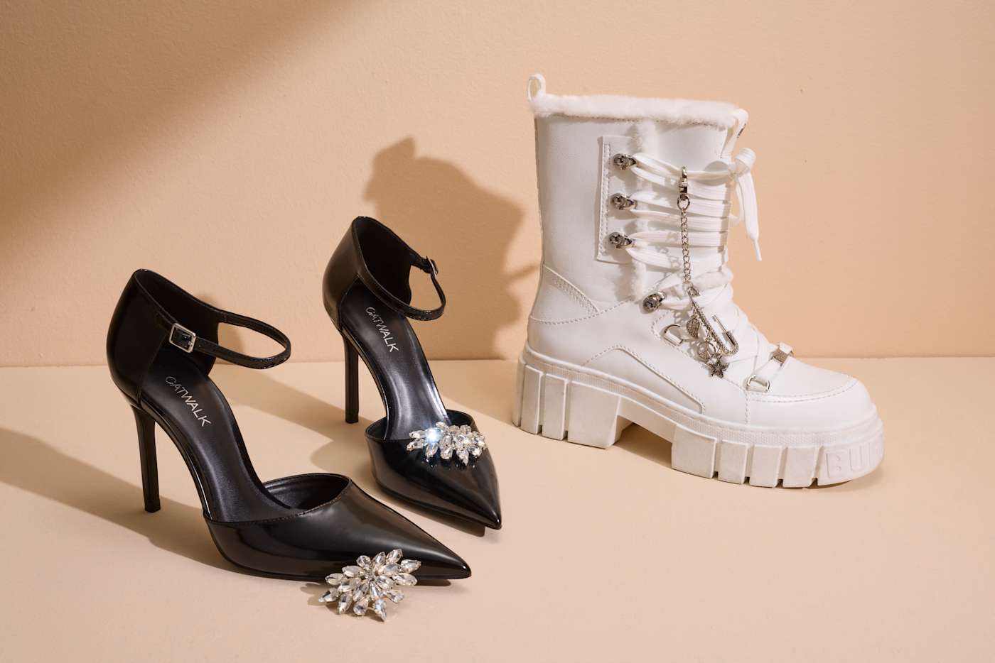 Zwei Paar Schuhe, elegante Pumps und Stiefel, mit dezenten Accessoires vor sandfarbenem Hintergrund