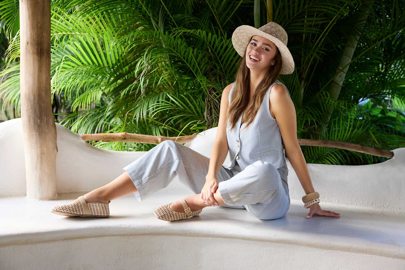 Frau in hellblauem Boho-Jumpsuit, sitzt auf Marmor-Bank, trägt Strohhut