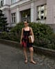 Frau in rotem Kleid, mit Handtasche unterm Arm, trägt offene Riemchen-Sandalen, stehend vor Hecke