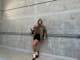 Frau in brauner Jacke, mit passendem Top und schwarzen Shorts, lehnt an Hauswand