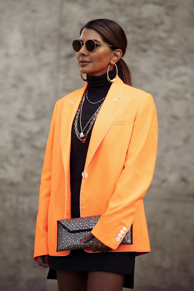 Stehende Frau in pfirsichfarbener Statement-Jacke, mit glitzernder Clutch in der Hand