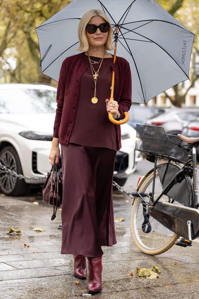Frau in elegantem Outfit in Bordeauxrot, lässig spazierend mit Schirm und Handtasche