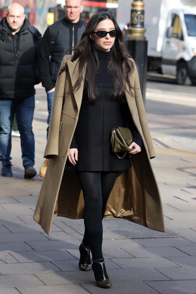 Mikey Madison trägt auf einem Gehweg einen langen beigefarbenen Mantel, schwarzen Rollkragenpullover, Minirock, Strumpfhosen und Stiefel, dazu Sonnenbrille und Handtasche.