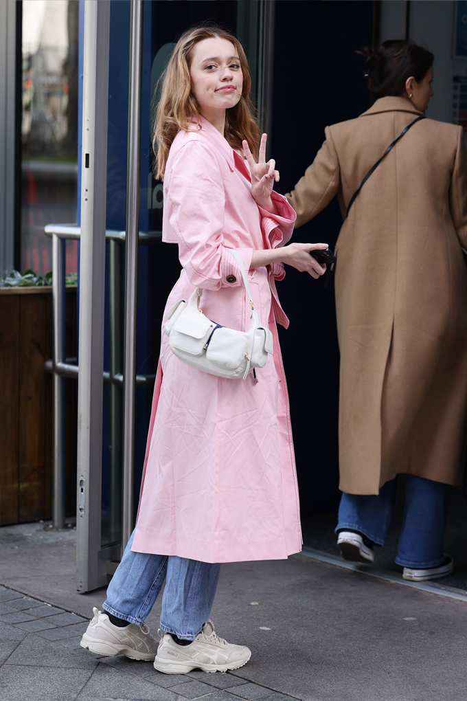 Aimee Lou Wood trägt Outfit mit pinkem Trenchcoat, kombiniert mit Denim-Hose und weißer Schultertasche