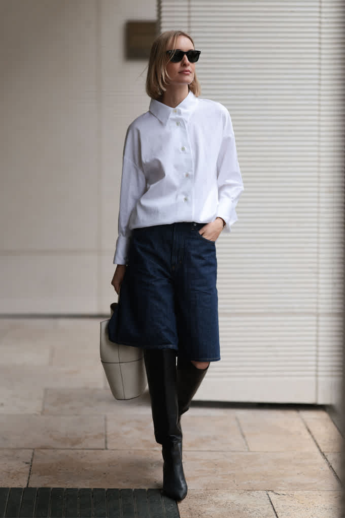 Frau trägt weiße Oversize-Bluse für den Office Siren-Look, kombiniert mit dunkelblauer Bermuda und Stiefeln