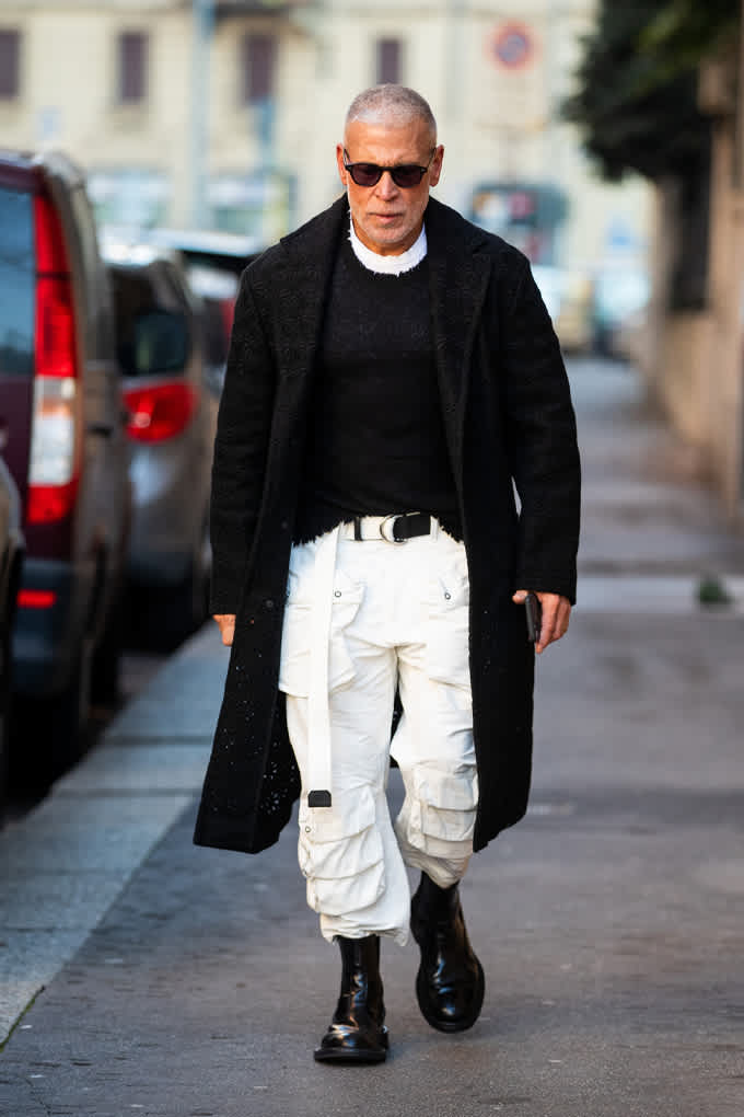 Mann mit schwarzem Strickpulli, passendem Longcoat und weißer Multi-Pocket-Pants