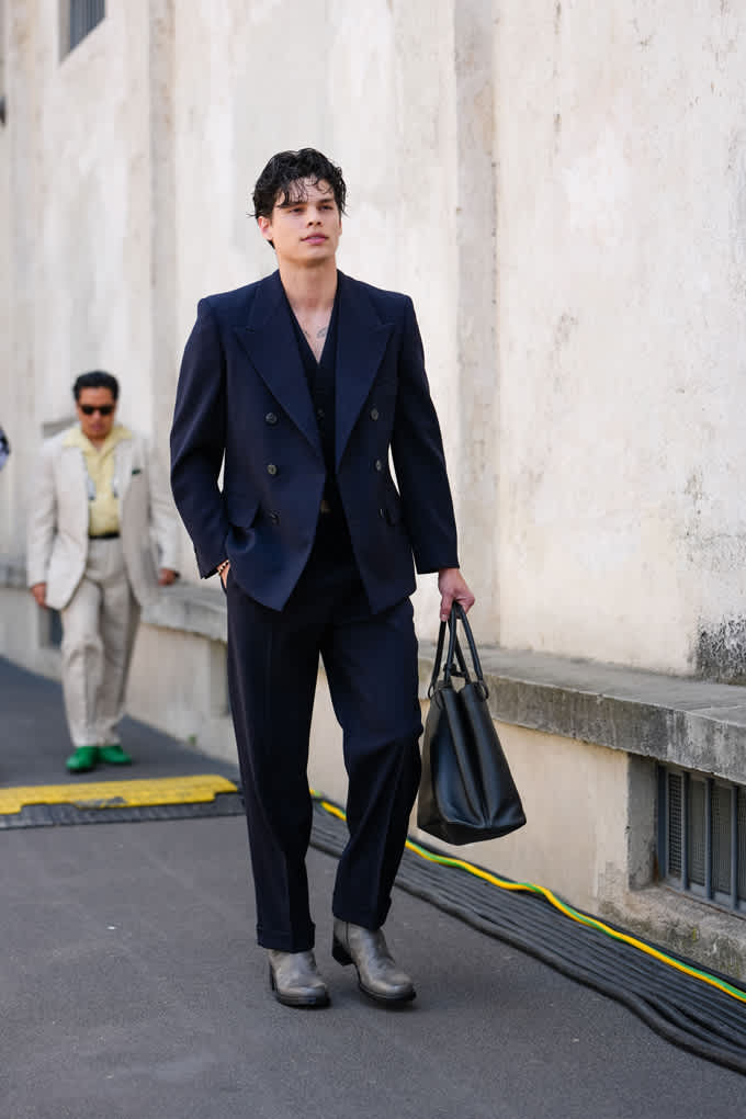Ein Mann trägt einen dunkelblauen Blazer, eine schwarze Anzugshose, graue Schuhe & hält eine schwarze Tasche in seiner Hand