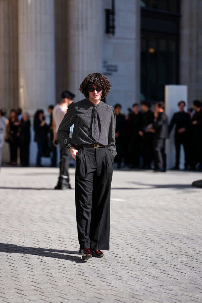Ein Mann mit dunkelbraunen Locken und einer Sonnenbrille trägt ein silbernes Hemd mit passender Krawatte und eine schwarze Anzugshose
