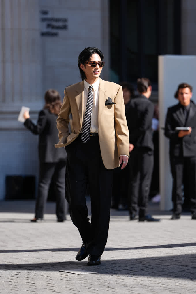 Ein Mann mit Sonnenbrille trägt einen beigen Blazer, ein weißes Hemd, eine gestreifte Krawatte und eine schwarze Anzugshose