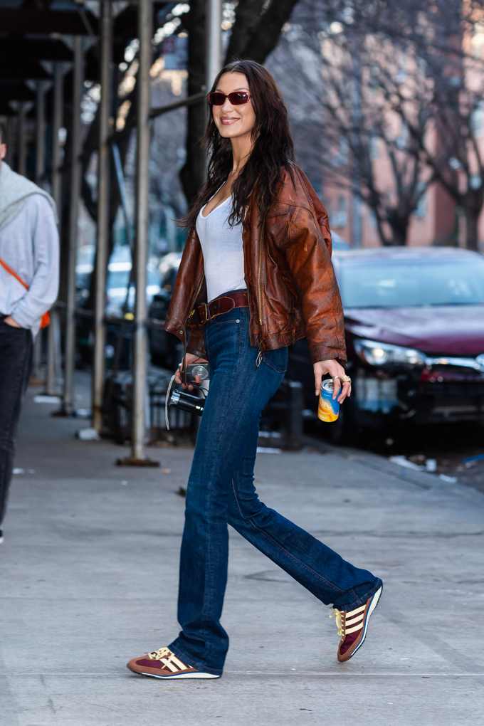 Eine Frau trägt eine braune Lederjacke, ein weißes Top, eine dunkelblaue Jeans und braune Sneaker
