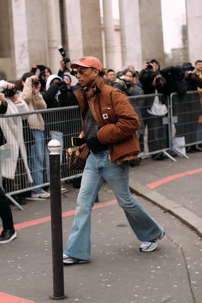 Ein Mann trägt eine braune Jacke, eine Schlagjeans, graue Sneakers und eine orange Cap