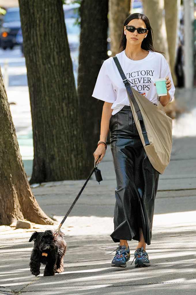 Eine Frau spaziert mit einem Hund und trägt ein weißes Print-T-Shirt, einen schwarzen Lederrock, bunte Sneakers und eine Umhängetasche