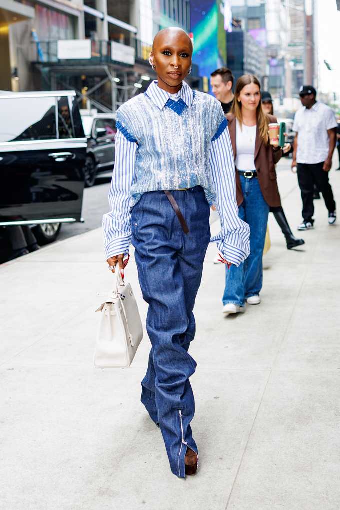 Cynthia Erivo trägt eine dunkelblaue Jeans, eines blau gestreiftes Hemd und einen hellblauen Pullunder, in der Hand trägt sie eine weiße Tasche