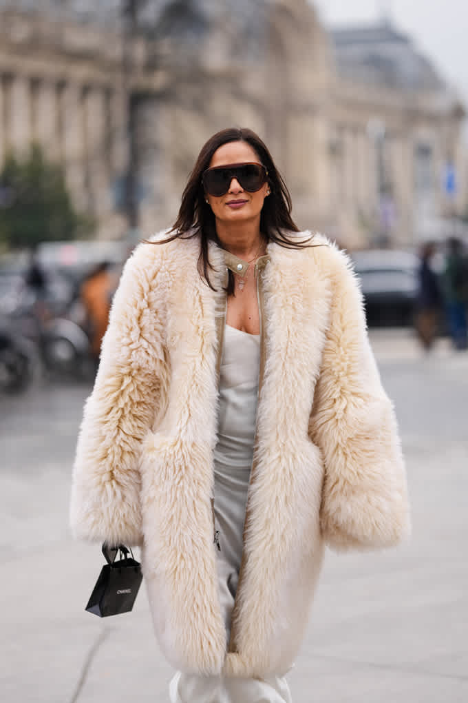 Eine Frau trägt einen voluminösen cremefarbenen Pelzmantel über einem eleganten, schimmernden Outfit und kombiniert ihn mit einer kleinen schwarzen Tasche und großer Sonnenbrille