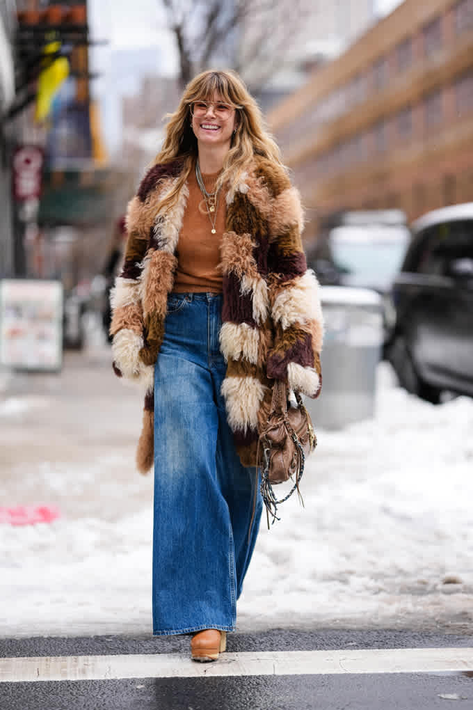 Die Frau trägt einen bunten Patchwork-Kunstpelzmantel zu weiter Jeans und braunem Pullover. Tasche und Schmuck ergänzen den lässigen Retro-Look