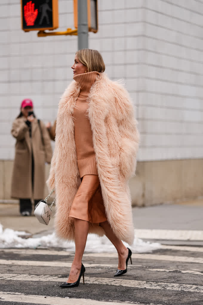 Eine Frau trägt einen flauschigen, roséfarbenen Mantel über einem Ton-in-Ton gehaltenen Rollkragenpullover und Midirock, dazu schwarze High Heels und eine weiße Tasche